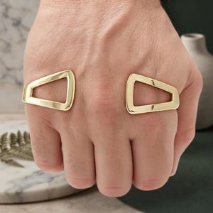 Può includere: Un bracciale rigido in metallo color oro con due estremità aperte di forma trapezoidale geometrica. La superficie lucida e liscia del metallo riflette la luce, mettendo in risalto le linee angolari e pulite di questo design aperto da polso.