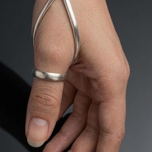 May include: A silver-toned hand ornament featuring a smooth, curved metal band that wraps around the wrist and extends to a circular ring encircling the thumb. The piece has a polished, metallic finish and a continuous, minimalist structural design.
