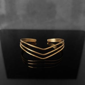 Puede incluir: Una pulsera tipo brazalete de tono dorado que presenta tres bandas paralelas que convergen en un punto central en forma de V o chevrón. El metal tiene un acabado liso y pulido con un diseño abierto para ajustar al contorno de la muñeca.