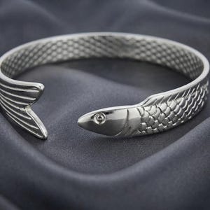 May include: A silver-toned metal cuff bracelet shaped like a fish. The bracelet features a textured scale pattern along the band, with a detailed fish head at one end and a fanned tail fin at the other, creating an open-ended circular design.