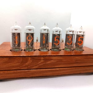 Könnte beinhalten: Eine digitale Holzuhr mit sechs Nixie-Röhren, die die Uhrzeit 19:53:55 anzeigt. Die Röhren haben orangefarbene Ziffern und sind in Glas eingeschlossen. Die Uhr steht auf einem Sockel aus gebeiztem Holz.