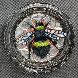 May include: A decorative glass coaster featuring a detailed bumblebee design. The bee has black and yellow stripes, with blue and white wings. The coaster has a silver rim and a textured background with red and gray accents.