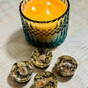 May include: A teal glass candle jar with a textured design, containing a lit, three-wick candle. Four resin coasters, each with embedded seashells and sand, are positioned below, evoking a seaside theme.