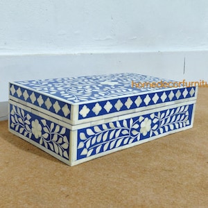 Pode incluir: Uma caixa decorativa retangular azul e branca com um padrão floral e geométrico. A caixa possui uma tampa articulada e é feita de um material de cor clara com incrustações azuis. A caixa tem aproximadamente 25 cm de comprimento.
