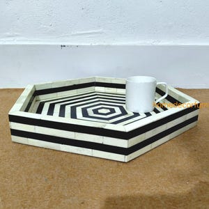 May include: A hexagonal tray with a black and white geometric design. The tray has a white border and a black and white striped pattern. A white mug sits on the tray. The tray is on a brown surface.