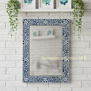 Puede incluir: Un espejo rectangular con un marco con estampado floral azul marino cuelga de una pared de ladrillos blancos. Encima del espejo hay una estantería blanca con tres pinturas de acuarela enmarcadas. Plantas verdes colgantes cuelgan a ambos lados del espejo. El texto "homedecorfurniture48" es visible.