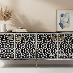 Puede incluir: Un aparador con estampado en blanco y negro con cuatro puertas, herrajes dorados y finas patas doradas. El mueble está decorado con un diseño floral. Un jarrón con flores secas, libros y obras de arte están encima.