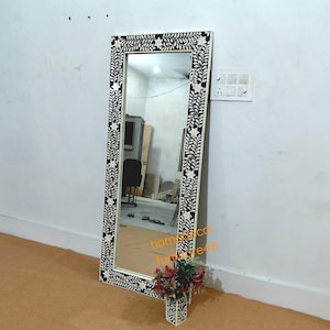 Puede incluir: Un espejo alto y rectangular con un marco decorativo. El marco presenta un patrón floral en blanco y negro. El espejo está apoyado contra una pared blanca. Un pequeño jarrón con flores está en la base del espejo.
