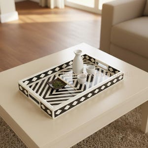 May include: A rectangular serving tray with a black and white geometric pattern, holding a white sake bottle, two small cups, and a dish with green contents. The tray sits on a cream-colored coffee table.