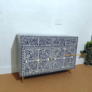 Op de afbeelding: Een marineblauwe en witte inlegkast met bloemmotief. De kast heeft twee deuren, vier laden en goudkleurige poten en hardware. De kast is een meubelstuk.