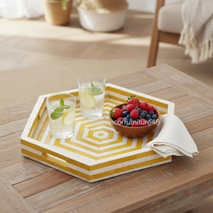 Peut inclure: Un plateau de service hexagonal avec un motif géométrique jaune et blanc. Il contient deux verres d'eau avec du citron et de la menthe, un bol en bois de baies et une serviette blanche pliée. Le plateau est sur une table en bois.