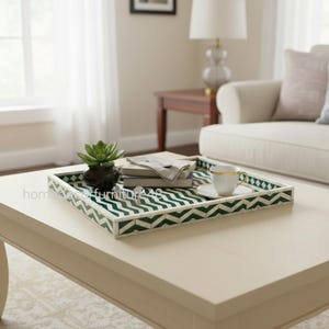 May include: A decorative rectangular tray with a green and white chevron pattern. The tray holds a small potted plant, books, a napkin, a teacup, and a saucer. The tray is on a cream-colored coffee table.