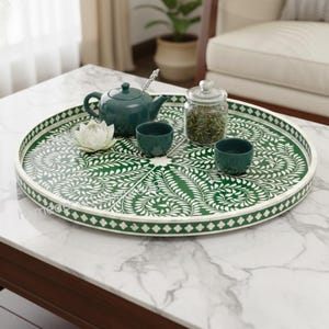 Puede incluir: Una bandeja redonda decorativa verde y blanca con un diseño floral, que contiene una tetera verde azulado, dos tazas pequeñas, un tarro de cristal y una flor blanca. La bandeja está sobre una mesa con tapa de mármol.
