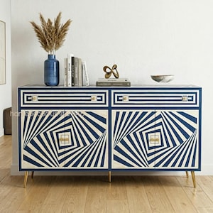 以下が含まれることがあります： 幾何学模様のネイビーブルーと白のキャビネット、金色の脚付き。キャビネットには2組のドアと引き出しがあります。乾燥した植物、本、装飾品が入った青い花瓶が上に置かれています。