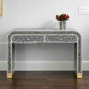 May include: A black and white bone inlay console table with two drawers. The table features a floral design and gold accents on the legs. A vase of red flowers sits on top of the table.