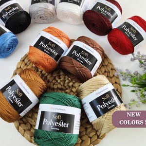 Puede incluir: Una colección de ovillos de hilo de colores, incluyendo azul, negro, blanco, rojo, naranja, marrón, verde y beige. Cada ovillo está etiquetado como "Soft Polyester" y descansa sobre una estera tejida. La imagen también incluye un letrero que dice "NEW COLORS !!".