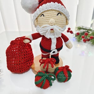 May include: A crocheted Santa Claus figure with a red suit, white beard, and hat. The figure holds a red sack and is surrounded by crocheted gift ornaments in red, green, and tan. A festive holiday decoration.