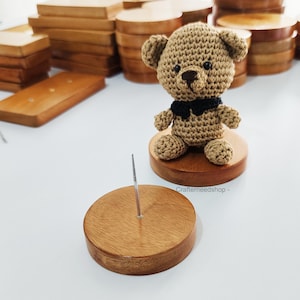 May include: A small crocheted tan teddy bear with a black bow tie sits on a round wooden base. Beside it is a matching round wooden base featuring a thin, upright metal spike. The wood has a smooth, polished finish with visible grain patterns.