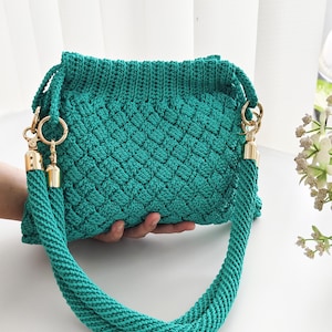 May include: A teal green crocheted handbag with a textured diamond pattern. The bag has matching rope handles with gold-colored hardware. The text "ANYAME" and "Crafterneedshop" are visible.