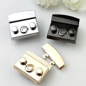 May include: Three metal purse clasps in silver, black, and gold. Each clasp has a rectangular base with three circular rivets and a central circular design. One clasp is open, revealing the hook mechanism.