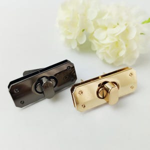 Puede incluir: Dos cierres metálicos para bolsos, uno en acabado cañón de fusil oscuro y otro en tono dorado brillante. Ambos tienen forma rectangular con un mecanismo de cierre central y remaches decorativos. Flores blancas en el fondo.