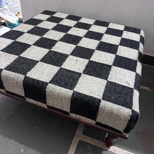 May include: A square ottoman featuring a woven fabric top with a black and white checkered pattern. The seat is supported by a dark brown wooden frame with short, turned legs that provide a stable base for the upholstered cushion.