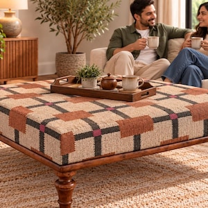 May include: A square ottoman featuring a textured fabric upholstery with a geometric grid pattern in shades of tan, brown, black, and small magenta accents. The piece is supported by four dark brown turned wooden legs and holds a wooden tray.