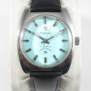 May include: A Tressa Laser Beam wristwatch with a light blue dial, silver-tone hour markers, and silver hands. The watch features a stainless steel case and a black leather strap. The dial displays the text TRESSA, 17 JEWELS, and LASER BEAM.