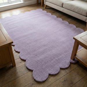 May include: A large, lavender-colored rug with a scalloped edge. The rug is rectangular with a soft, textured surface. It is placed on a hardwood floor, with wooden side tables and a sofa visible in the background.