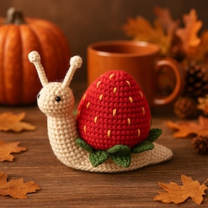May include: A handmade crochet snail with a strawberry shell. The snail's body is cream-colored, and the strawberry shell is red with yellow seed details. Green leaves are at the base of the strawberry. The snail is on a wooden surface, with a pumpkin and mug in the background.