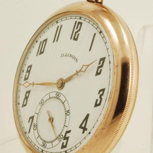 Könnte beinhalten: Eine goldfarbene Taschenuhr mit weißem Zifferblatt und schwarzen Ziffern. Das Zifferblatt hat ein kleineres Hilfszifferblatt und das Wort "ILLINOIS" ist darauf gedruckt. Die Uhr hat goldfarbene Zeiger und eine Schlaufe oben.