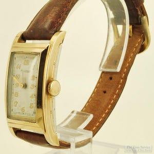 Può includere: Un orologio da polso vintage color oro con quadrante rettangolare e cinturino in pelle marrone. Il quadrante mostra numeri e lancette. Il cinturino è cucito con filo color chiaro e ha una fibbia color oro.