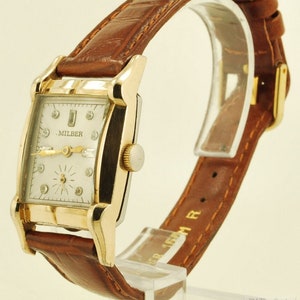 May include: A gold-tone wristwatch with a square face and a brown leather strap. The watch face has a white background with black Roman numeral hour markers and a diamond-shaped minute hand. The watch face also has the text ".MILBER" printed on it.