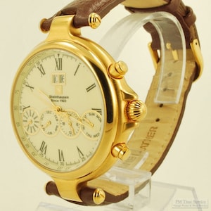 May include: A gold-tone wristwatch with a white face and Roman numeral hour markers. The watch has a brown leather strap and features a chronograph, date, and day of the week display. The watch face also includes the text "Steinhausen Since 1923".