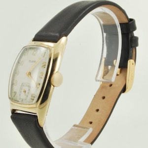 Op de afbeelding: Een gouden polshorloge met een witte wijzerplaat en een zwarte leren band. De wijzerplaat van het horloge heeft zwarte Romeinse cijfers als uurmarkeringen en een kleine subdial op 6 uur. Het horloge is gemarkeerd met "ELGIN" op 12 uur.