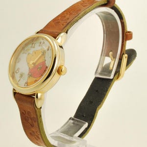 May include: A gold-tone wristwatch with a brown leather band. The watch face features a cartoon image of Winnie the Pooh. The band has a paw print design. The watch is displayed on a clear stand.