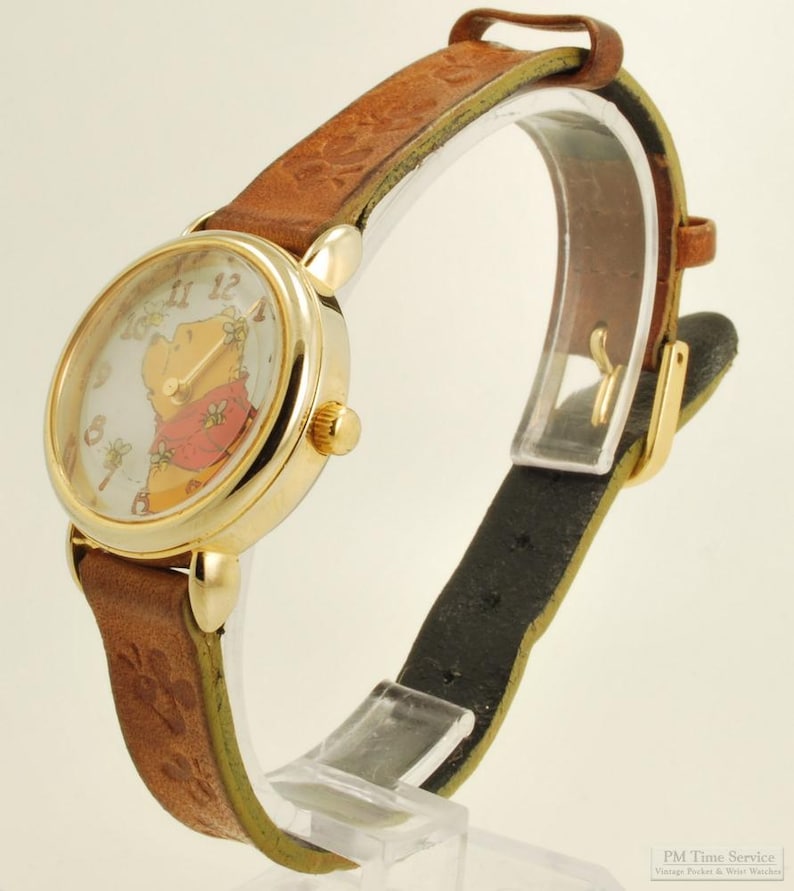 May include: A gold-tone wristwatch with a brown leather band. The watch face features a cartoon image of Winnie the Pooh. The band has a paw print design. The watch is displayed on a clear stand.