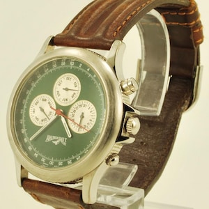 Könnte beinhalten: Eine silberfarbene Armbanduhr mit grünem Zifferblatt und braunem Lederarmband. Das Zifferblatt hat drei Hilfszifferblätter, eine Datumsanzeige und den Text "AMERICAN EAGLE". Das braune Lederarmband ist mit einem helleren Faden genäht.