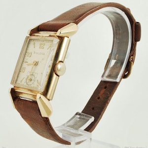 May include: A vintage Bulova wristwatch with a rectangular gold-tone case and a cream dial. It has gold hands and markers, and a brown leather strap. The watch is displayed on a clear stand. The watch strap is brown.