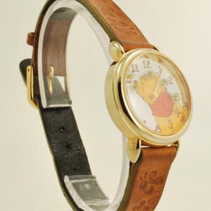 May include: A gold-tone wristwatch with a brown leather band. The watch face features a cartoon character, and the band has a floral pattern. The watch has a black leather backing and a gold buckle. The watch is displayed on a clear stand.