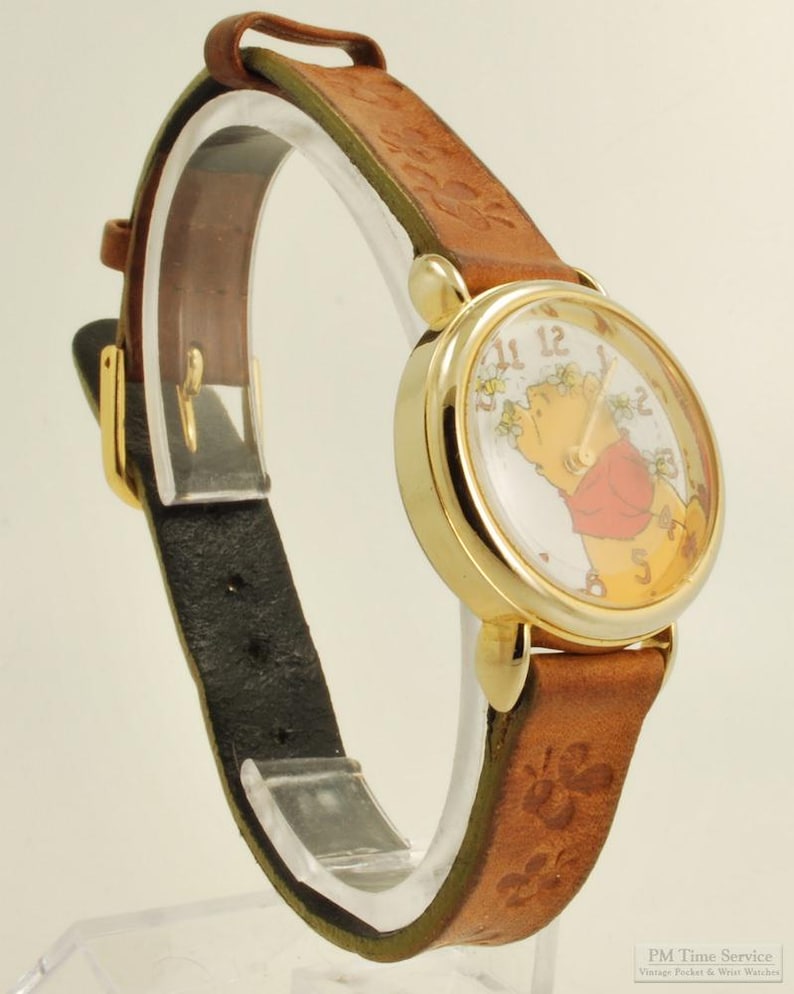 May include: A gold-tone wristwatch with a brown leather band. The watch face features a cartoon character, and the band has a floral pattern. The watch has a black leather backing and a gold buckle. The watch is displayed on a clear stand.