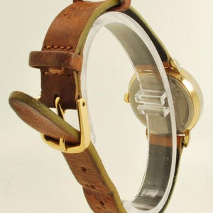 May include: A vintage gold-toned wristwatch with a brown leather band. The watch has a round face and a gold buckle. The leather band is stitched and shows signs of wear. The watch is displayed on a clear stand.