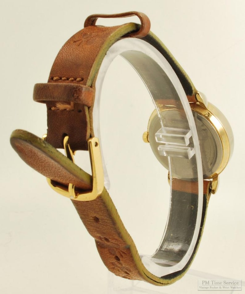 May include: A vintage gold-toned wristwatch with a brown leather band. The watch has a round face and a gold buckle. The leather band is stitched and shows signs of wear. The watch is displayed on a clear stand.