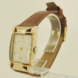 Könnte beinhalten: Eine Vintage-Bulova-Uhr mit einem rechteckigen, goldfarbenen Gehäuse und einem hellen Zifferblatt. Die Uhr hat goldene Zeiger und Stundenmarkierungen, und der Markenname ist sichtbar. Sie hat ein braunes Lederarmband.