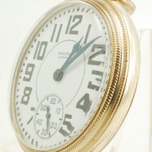 May include: A gold-toned pocket watch with a white face and black numerals. The watch has a subdial for seconds and blue hands. The text "WALTH 21 JEWELS" is visible on the face. The watch is a vintage pocket watch.