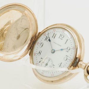 May include: A gold-toned Waltham pocket watch with an open lid. The watch face is white with black numerals and hands, and a smaller subdial. The watch is open, revealing the inner workings and a second lid.