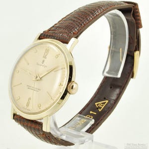 May include: A gold Sovereign wristwatch with a cream-coloured face and brown leather strap. The watch face displays the number 12, and the words "Shock Resistant Water Proof". The strap has a textured pattern.