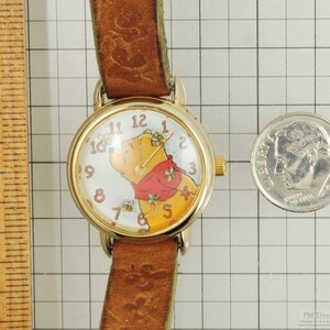 May include: A gold-toned wristwatch with a brown leather band. The watch face features a Winnie the Pooh design with a red scarf and bees. A U.S. dime is next to the watch for size comparison.