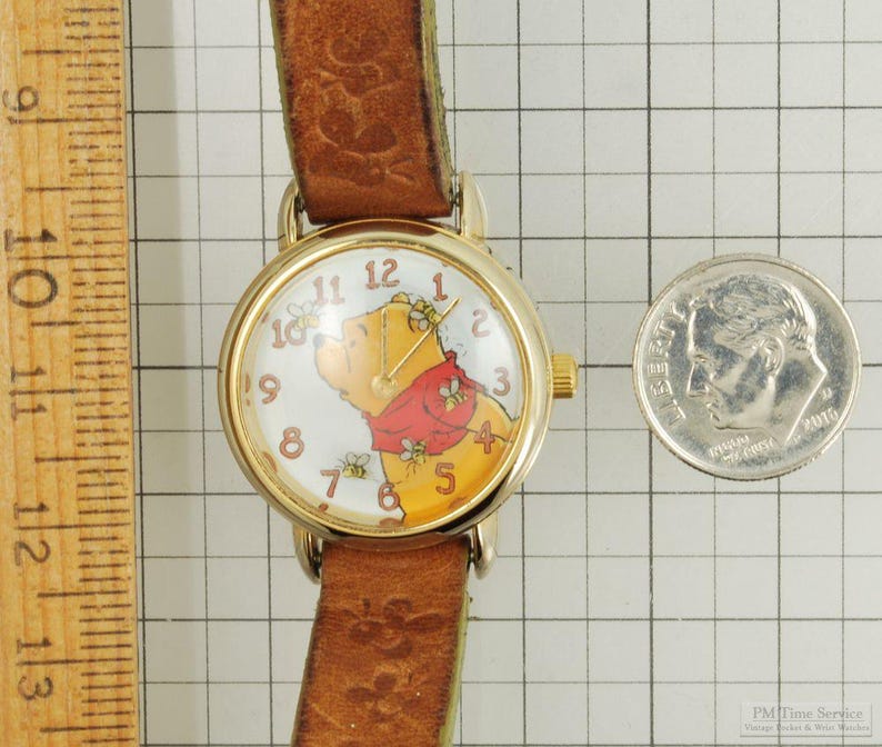 May include: A gold-toned wristwatch with a brown leather band. The watch face features a Winnie the Pooh design with a red scarf and bees. A U.S. dime is next to the watch for size comparison.
