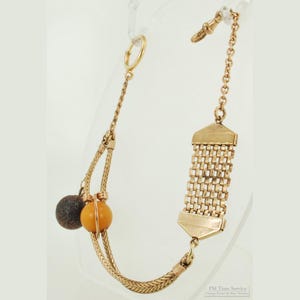 May include: A gold-toned watch chain with a rectangular woven section and a chain with a clasp. The chain has a loop on one end and two decorative spheres, one brown and one orange, on the other end. The chain is likely vintage.